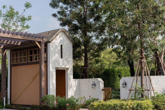 A white brick structure with a timber door under a pergola, beside a garden with wooden plant frames