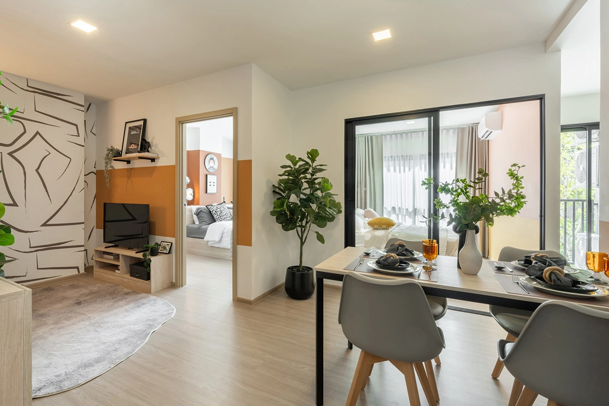 The living area of a two-bedroom: a television on a low unit against the orange wall, the door to the first bedroom on the left, the second bedroom behind the sliding glass panel on the right and a dining table for four by the window