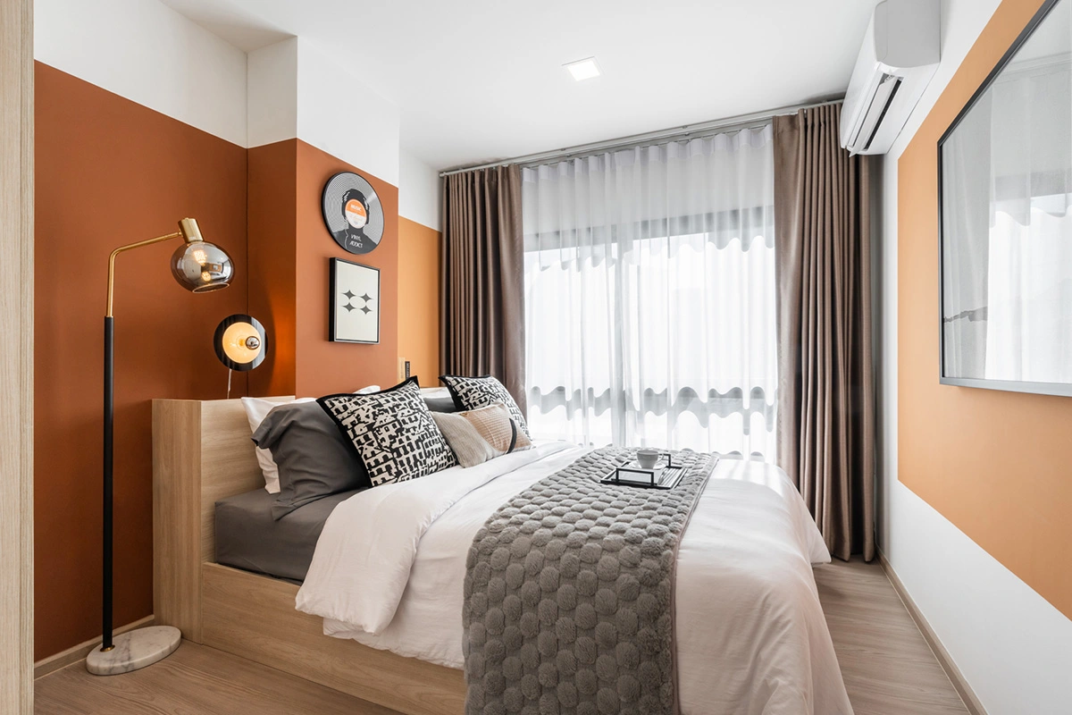 A show-unit bedroom: a double bed with a timber headboard against a terracotta wall, a floor lamp and a full-height window with double curtains