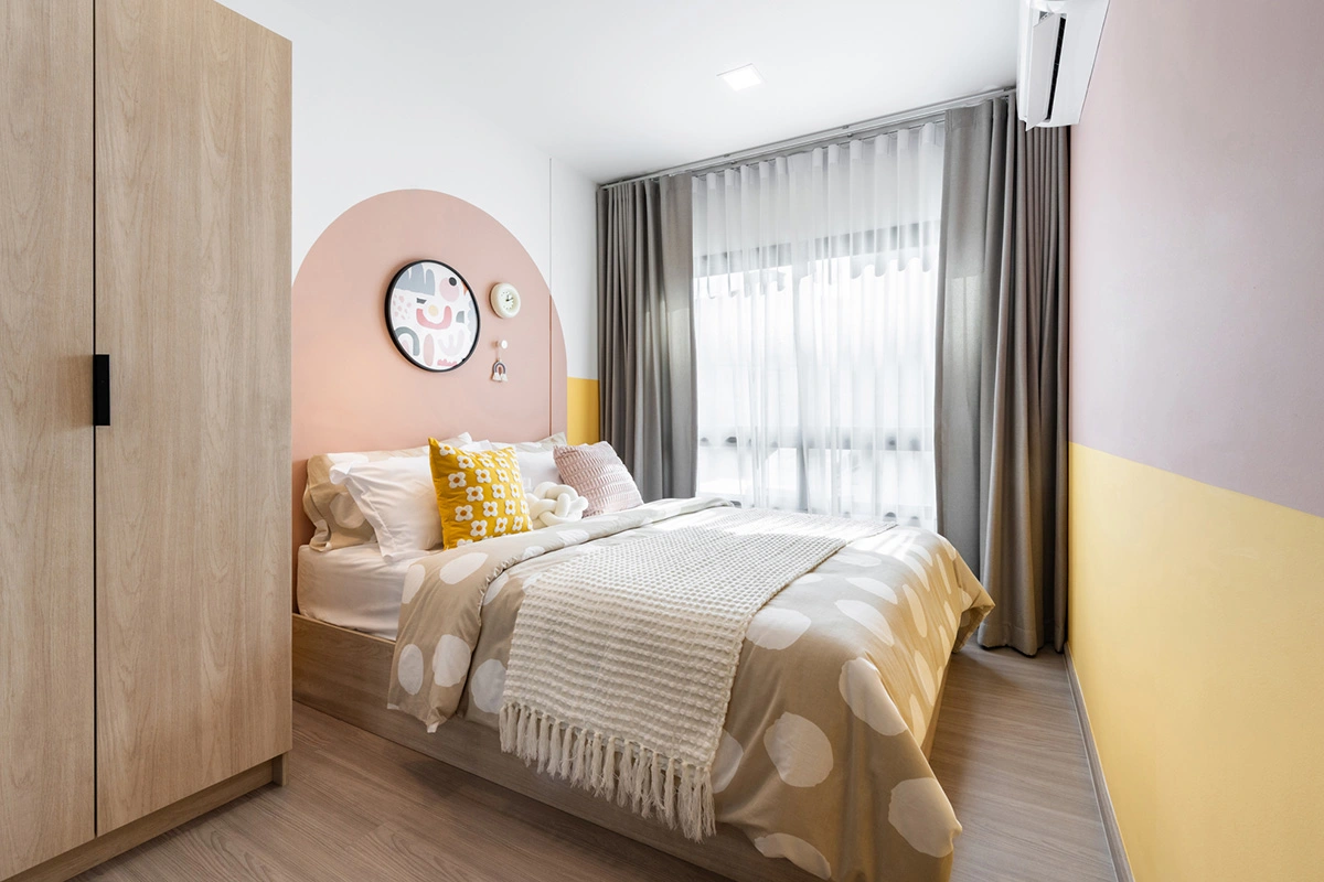A bedroom in pink and yellow: a full-height wardrobe on the left, the bed against an arched headboard panel and a window with grey curtains