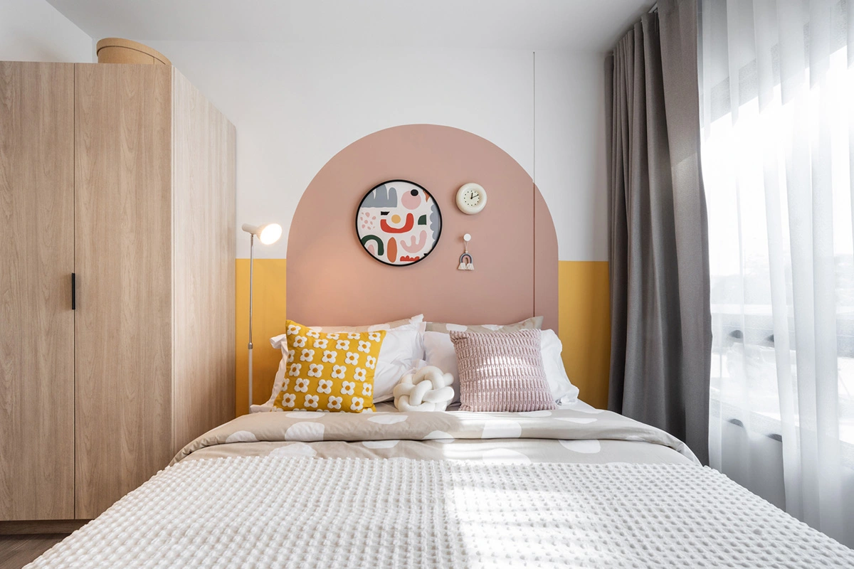 The same bedroom head-on: the painted arch with a round picture and a clock, cushions on the bed, the wardrobe on the left and sheer curtains on the right