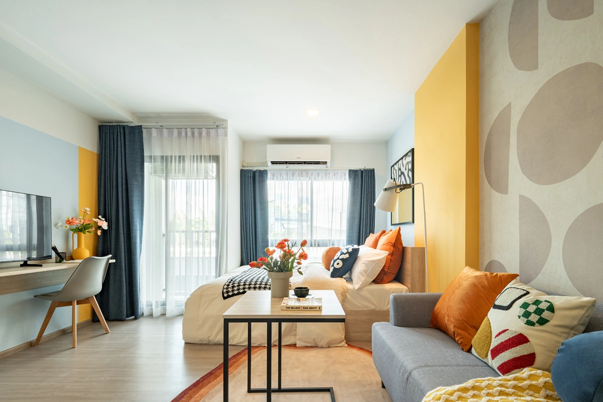 The room in full: bed and sofa in one volume with no partition, the working table with a television by the wall, the balcony door and a window with blue curtains