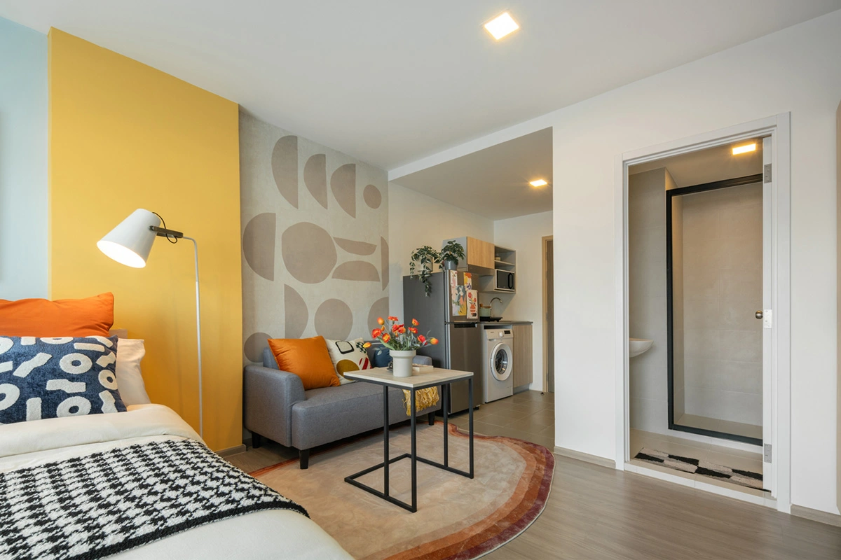 The same room from the bed: sofa and side table on the rug, the kitchen with fridge and washing machine beyond, and the bathroom door on the right