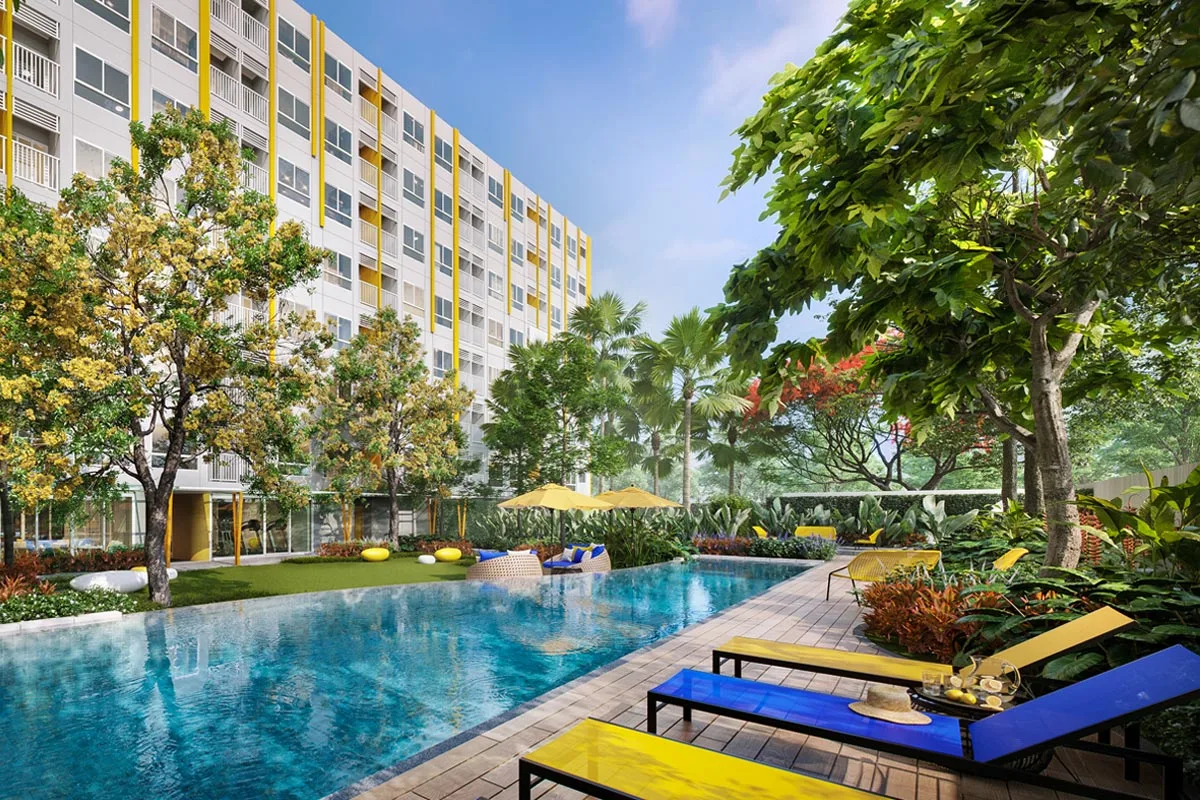 The pool along the building: yellow and blue loungers, a lawn, a communal room behind the ground-floor glass, palms and flowering trees around
