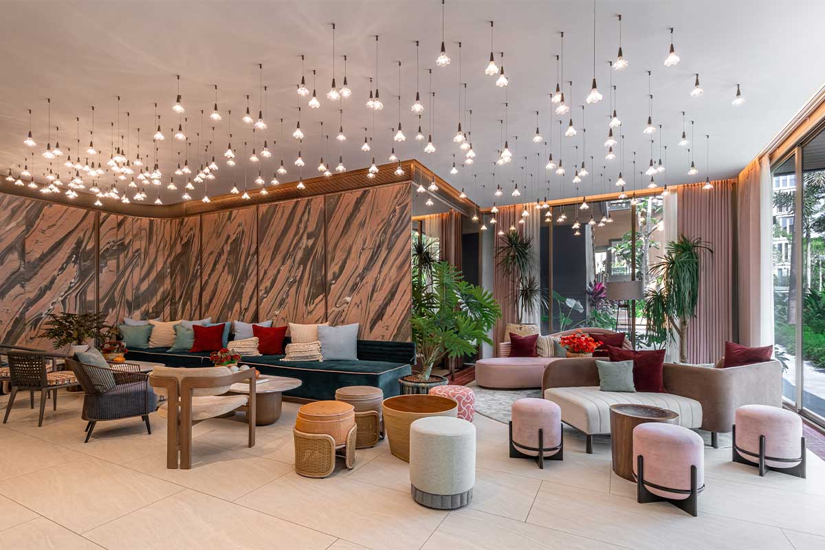 The lobby: a wall of pink-and-brown marble, bulbs on cords of differing lengths across the whole ceiling, a green sofa, powder-pink poufs and full-height windows onto greenery