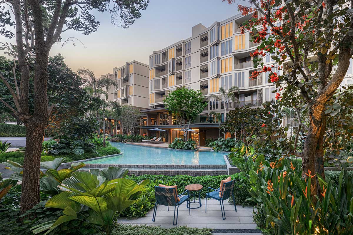 The pool at dusk: water running along the building, loungers by the glazed communal area, a table and chairs in the garden in the foreground and the second building on the left