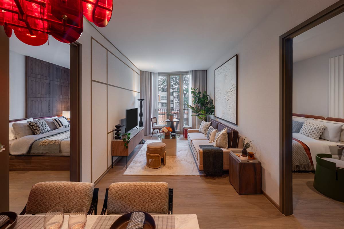 A two-bedroom show unit: the living room with sofa and television between the two bedrooms, beds visible through the openings on either side, and at the end floor-to-ceiling glazing with the railing flush against the glass, a table and chair standing in the room in front of it