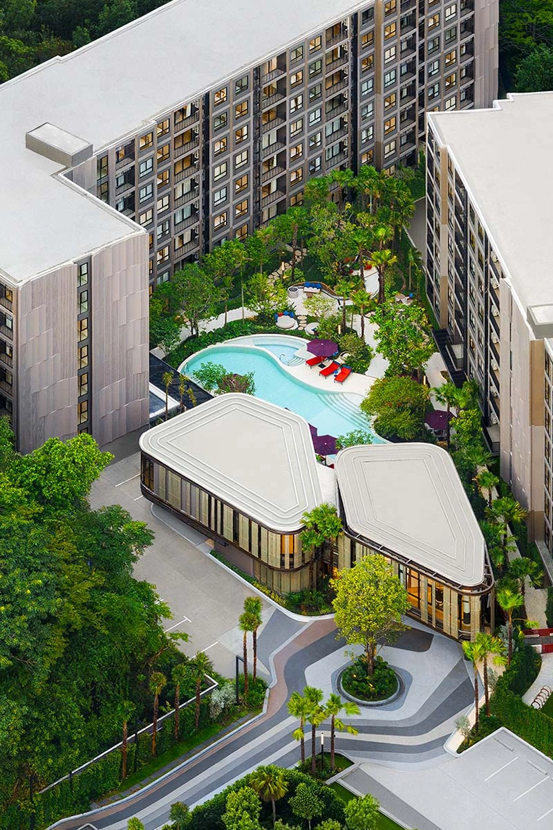Straight-down view by day: two pavilions with white faceted roofs, the pool with orange loungers and purple parasols behind them, garden and parking along the buildings