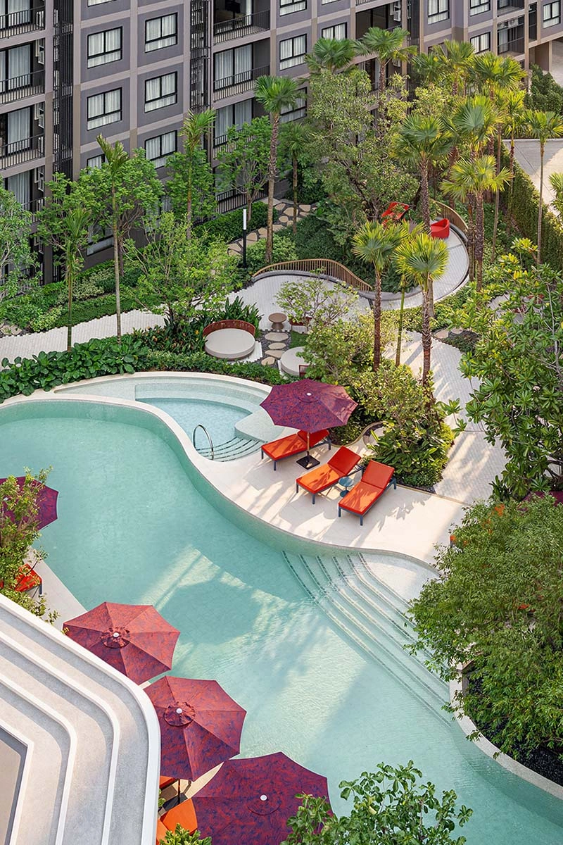 The pool from above by day: a curved basin, purple parasols and orange loungers along its edge, palms and dense garden between the wings