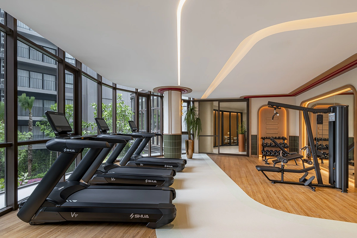 The fitness room: treadmills along the full glazing, a weights machine and a dumbbell rack further in, a timber floor and a wavy ceiling