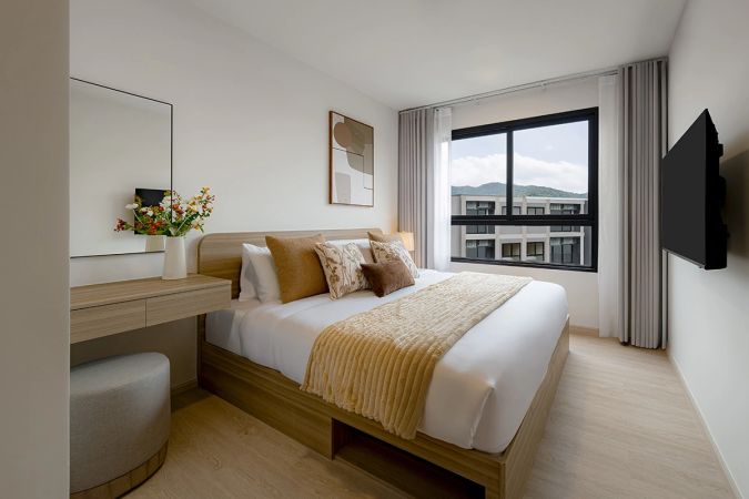 A bedroom: a bed with a mustard throw, a dressing table with a mirror and a pouf, a wall-mounted TV and a window looking at the hills