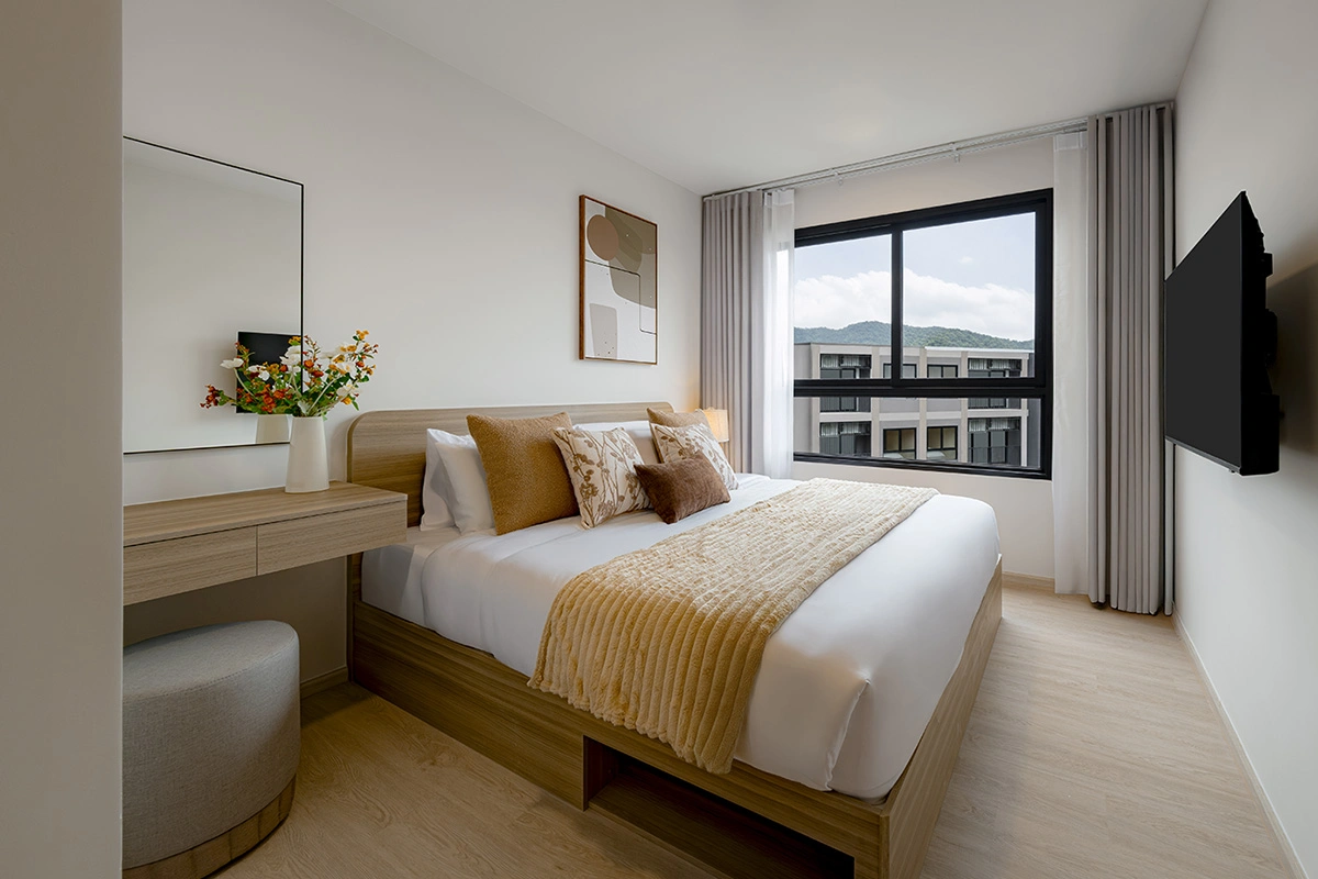 A bedroom: a bed with a mustard throw, a dressing table with a mirror and a pouf, a wall-mounted TV and a window looking at the hills