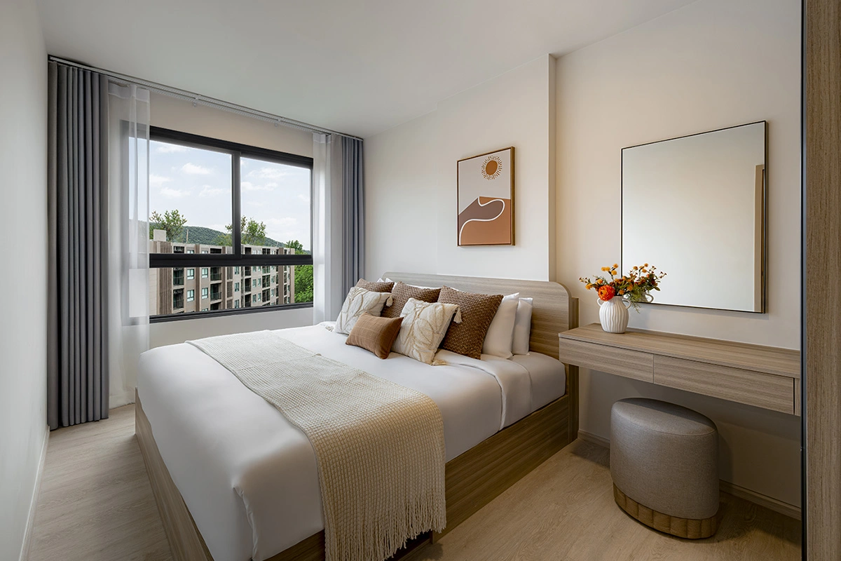 A bedroom from another angle: the bed against the wall, a dressing table with a square mirror, a round pouf and a window onto the neighbouring building and the hills