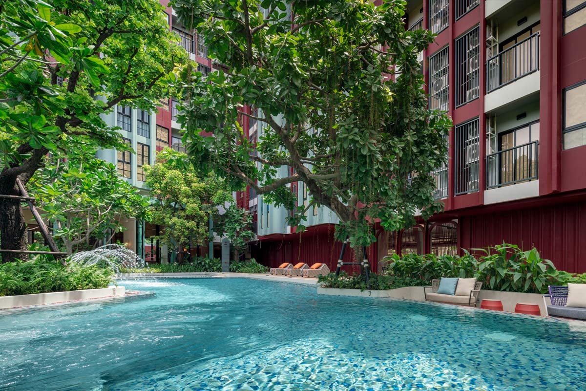 The pool from the far end: fountain jets on the left, a tree standing right over the water, three loungers with orange mattresses along the far edge and, closer in, a cushioned sofa, a dark-blue daybed and red stools at the water