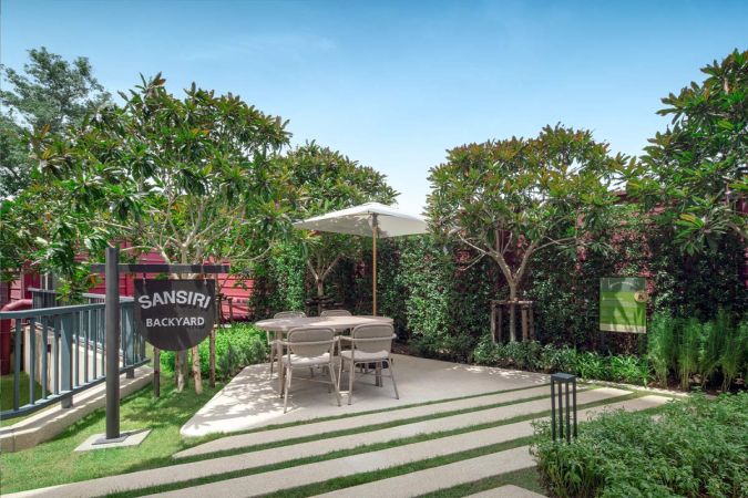 Sansiri Backyard: the name board, a dining table under a parasol on the paved area, frangipani trees and a hedge