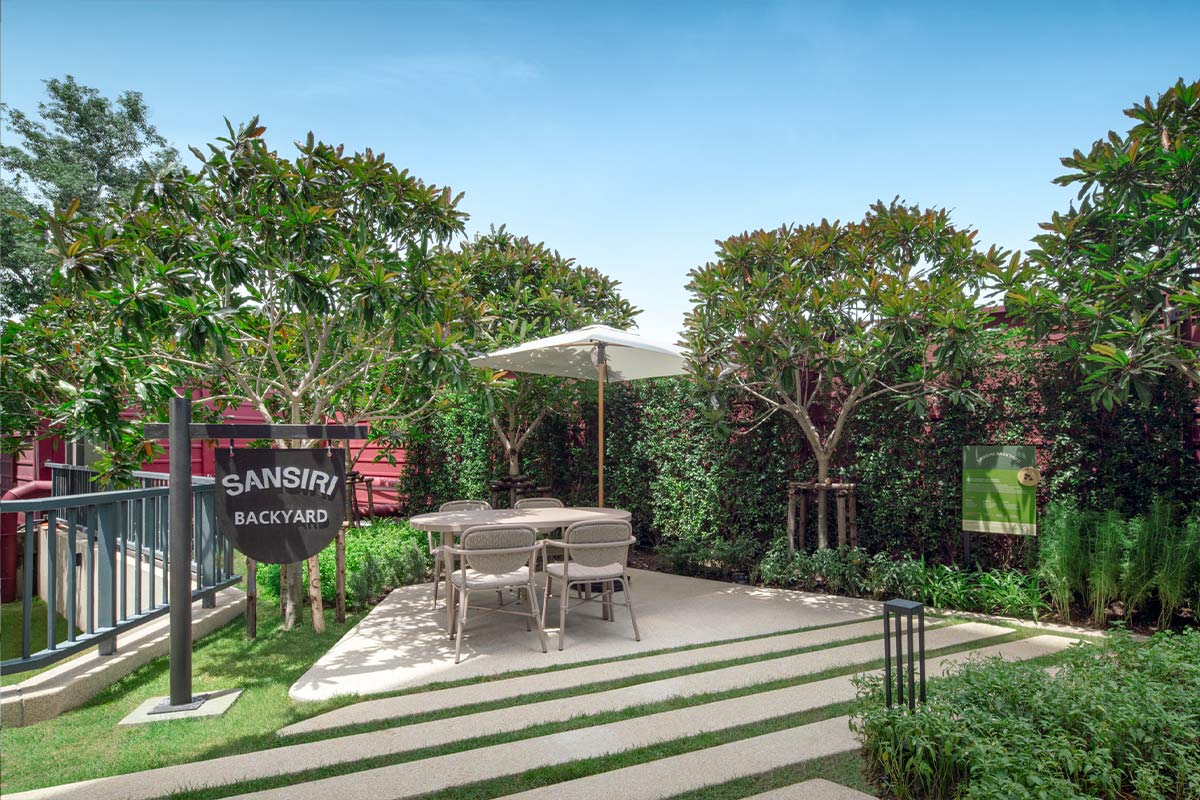 Sansiri Backyard: the name board, a dining table under a parasol on the paved area, frangipani trees and a hedge