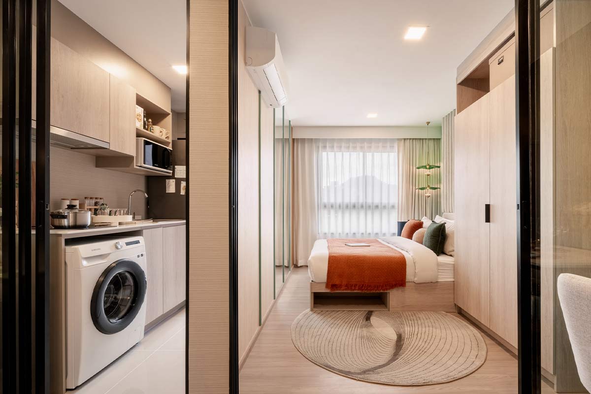 The view from the entrance into the unit: on the left the kitchen behind the black frame of a glass partition, then the bed with an orange throw by the window, the outline of a hill showing through the sheer curtain