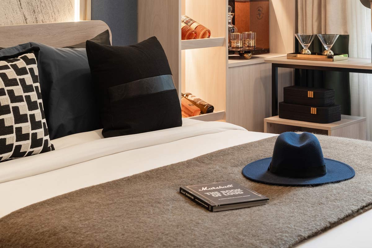 A close-up of the bed in a dark scheme: black and graphic cushions, a hat and a book on the throw, a shelf with glassware behind the head