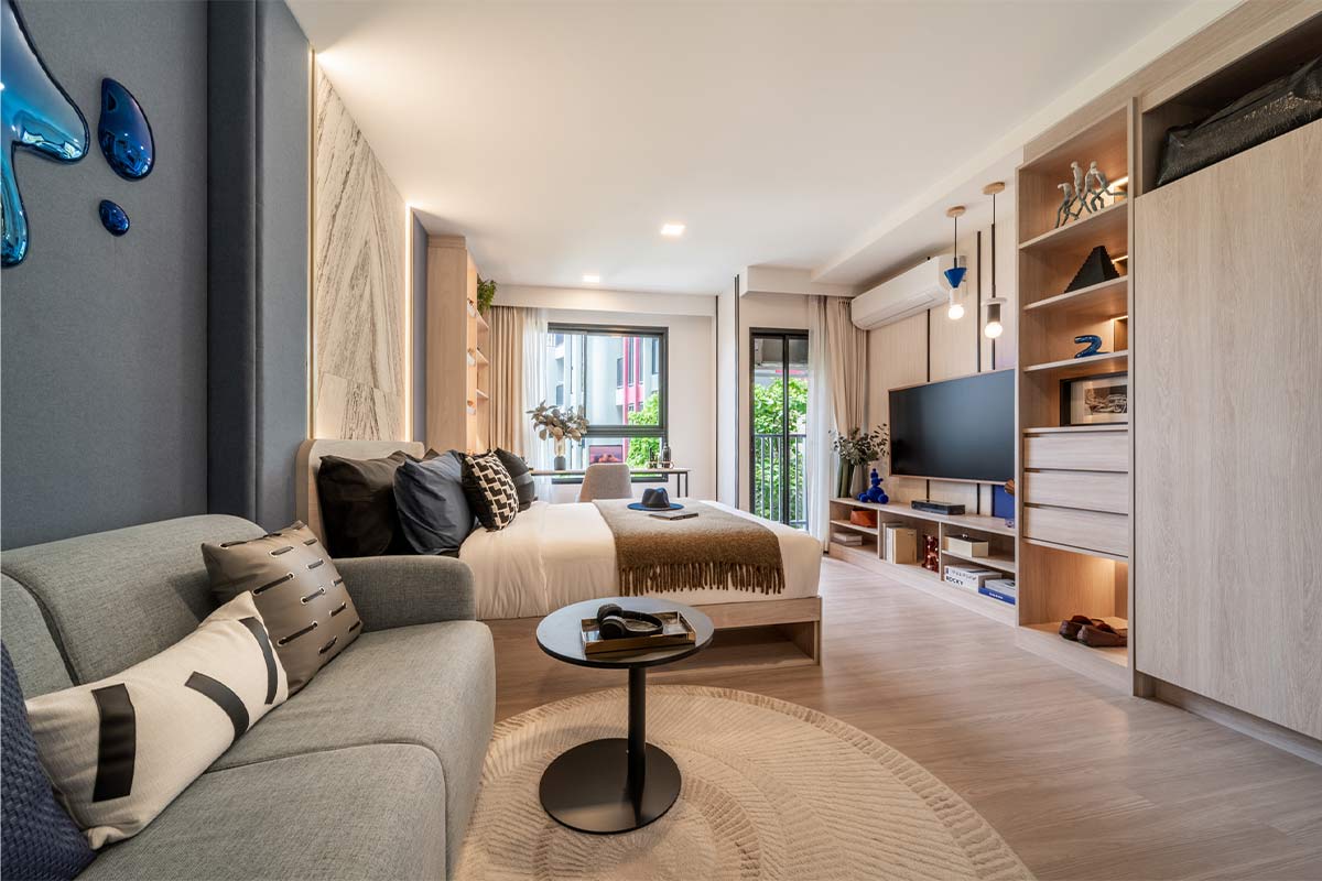 The same unit seen wider: the sofa in the foreground, the bed in the middle, the television on a cabinet and the balcony door on the right